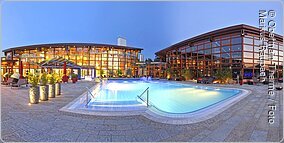 Bad Staffelstein, Vitalbecken der Obermain Therme Zwei Gebäude mit breiter Glasfront, davor ein blau leuchtendes Wasserbecken